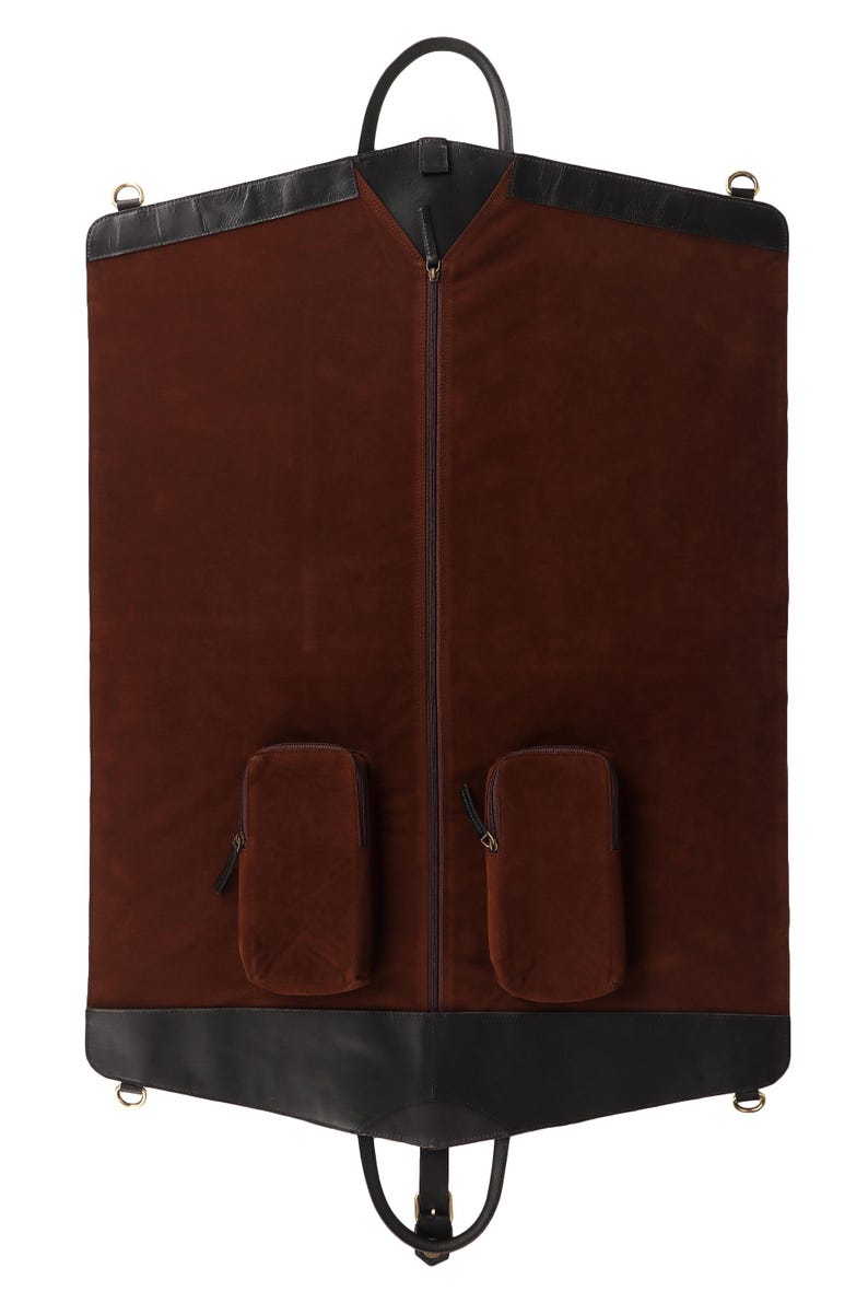 May include: A brown garment bag with black leather trim and a handle. The bag has a full-length zipper and two small pockets. The bag is designed to protect clothing during travel.
