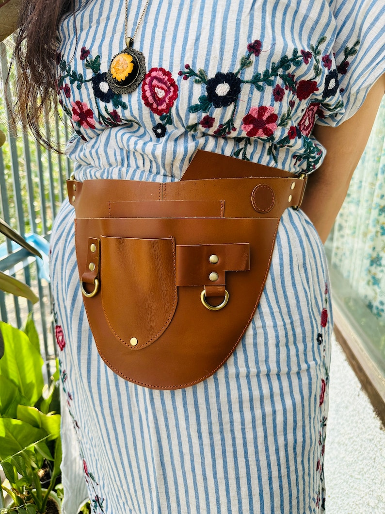 May include: A brown leather tool belt with a large pocket and multiple smaller pockets. The belt is worn over a blue and white striped dress with floral embroidery.