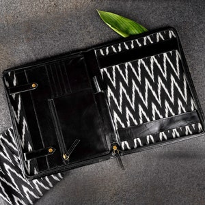 May include: Black leather portfolio with a white and black zigzag patterned interior. The portfolio is open, revealing multiple compartments, a pen holder, and a zippered closure. Gold-toned hardware accents the design. A green leaf rests inside.