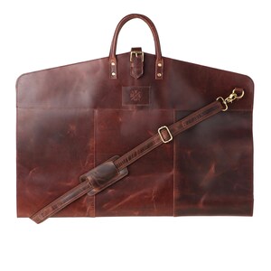 May include: A brown leather garment bag with a handle, buckle, and detachable shoulder strap. The bag has a rectangular shape with a curved top and a small logo. The bag is designed to carry suits and other clothing.