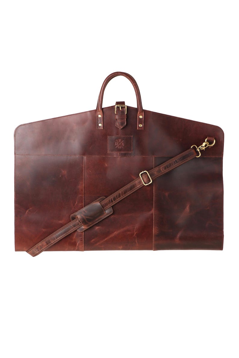 May include: A brown leather garment bag with a handle, buckle, and detachable shoulder strap. The bag has a rectangular shape with a curved top and a small logo. The bag is designed to carry suits and other clothing.