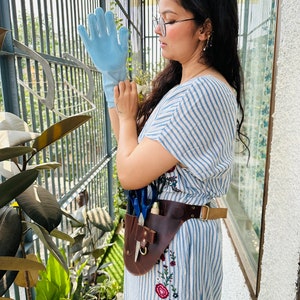 Puede incluir: Un guante de limpieza de silicona azul claro con cerdas texturizadas en la palma y los dedos, junto a un cintur&oacute;n utilitario de cuero marr&oacute;n oscuro con m&uacute;ltiples bolsillos y herrajes met&aacute;licos, sobre un vestido de rayas blancas y azules bordado.