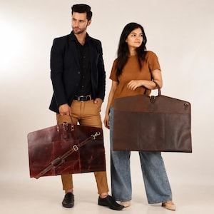 May include: Two rectangular leather garment bags in shades of dark brown and reddish-brown. Each bag features a top handle, a central buckle closure, and a detachable shoulder strap with metal hardware, designed for carrying suits or formal attire.