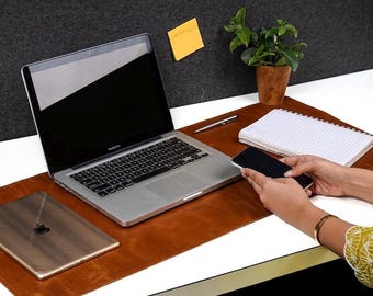 May include: A brown leather desk mat featuring a silver laptop, a tablet, a spiral-bound notebook, and a silver pen. The mat provides a smooth surface for office equipment, creating a workspace setup with a small potted plant nearby.