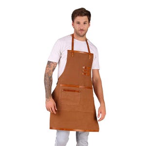 Könnte beinhalten: Eine braune Schürze mit Lederakzenten, einschließlich Riemen, Taschenbesatz und dekorativen Nieten. Die Schürze hat eine große Fronttasche und wird von einer Person in einem weißen T-Shirt getragen.