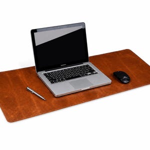 Può includere: Un sottomano rettangolare in pelle marrone con un computer portatile argentato, un mouse nero e una penna argentata posizionati sulla superficie. Il tappetino presenta angoli arrotondati e una consistenza liscia e uniforme su tutta l'area.