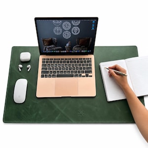 Peut inclure: Un sous-main en cuir vert foncé avec un logo subtilement gaufré, supportant un ordinateur portable rose doré, une souris sans fil blanche, des écouteurs blancs dans leur boîtier et un carnet ouvert à pages lignées avec un stylo argenté.