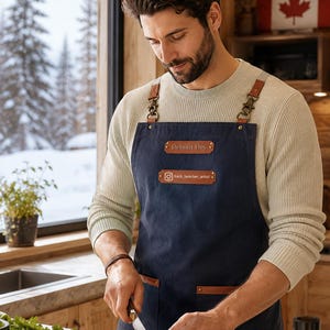 May include: A navy blue apron featuring brown leather straps with brass-toned metal clips, two brown leather rectangular patches, and two front pockets. The top patch displays the text Débolit Dey, and the lower patch shows an Instagram icon with the text back_bencher_artist.