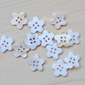 May include: A collection of 11 white, pearl-like, flower-shaped buttons with five points. Each button has four holes in the centre.