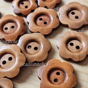 May include: A collection of flower-shaped wooden buttons in a warm brown hue. Each button features a central recessed area with two small holes. The buttons are scattered on a light-coloured wooden surface. The text "Avocadostitches" is visible.