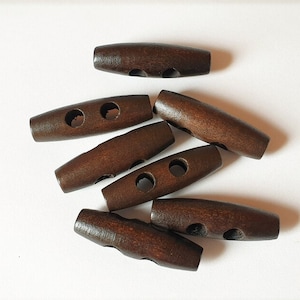 May include: A group of six brown wooden toggle buttons with two holes each. The buttons are arranged in a loose pile on a white surface.