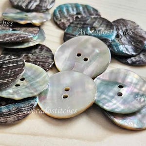 May include: A selection of iridescent, round buttons, each with two holes. The buttons display natural, organic patterns in shades of grey, brown, and teal. They are arranged on a light-coloured wooden surface, suitable for craft or sewing.