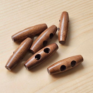 May include: Six wooden toggle buttons in a warm brown colour are arranged on a light wood surface. Each button is oblong with two or three holes for sewing. The buttons have a smooth, polished finish, suggesting they are new.