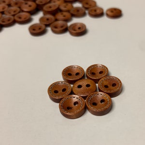 May include: A close-up of seven brown wooden buttons with two holes and a white stitched border.