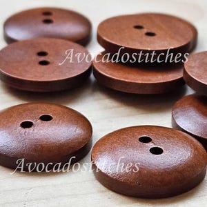 May include: A collection of large, circular, brown wooden buttons, each with two holes. The buttons have a smooth, polished appearance and are arranged on a light surface. The text "Avocadostitches" is printed on some of the buttons.