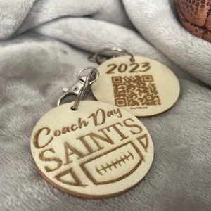 Pode incluir: Dois chaveiros de madeira com acabamento em madeira marrom. Um chaveiro tem o texto "Coach Day Saints" com um gráfico de futebol. O outro chaveiro tem o ano "2023" e um código QR.