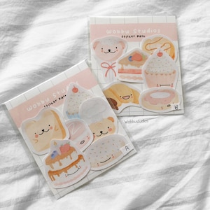 May include: Two sticker packs with adorable food and animal illustrations. One pack includes a bear, cake, cupcake, and donut. The other pack has bread, cake, a bear, and a sprinkle-covered pastry. The text "Wobbu Studios" is visible.