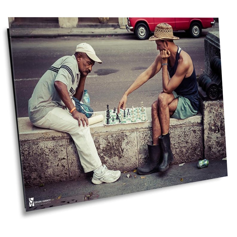 May include: Two men play chess on a street in Cuba. One man is wearing a white hat and the other is wearing a straw hat. They are sitting on a stone wall.