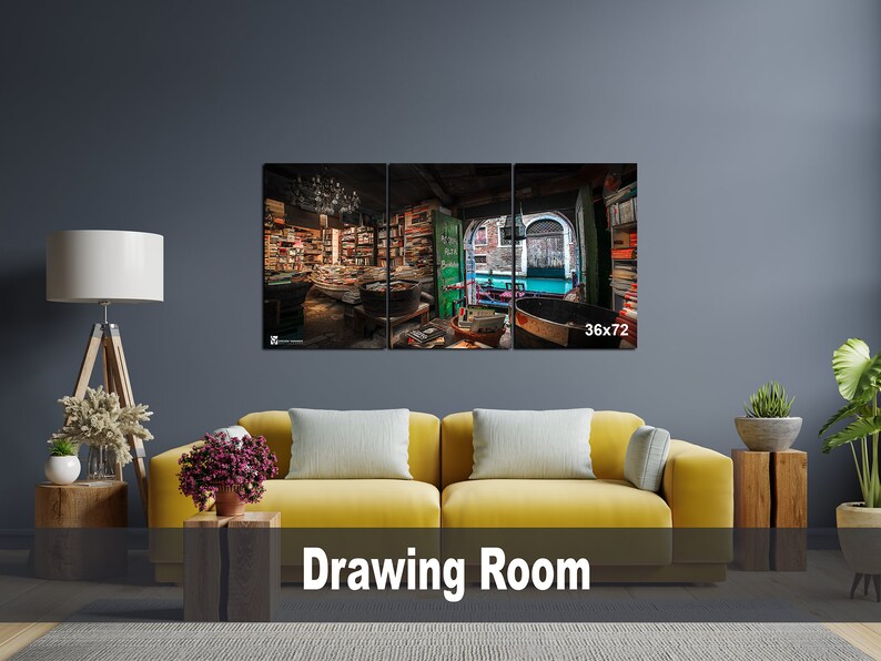 May include: A three-panel canvas print depicting a bookstore interior with bookshelves filled with books. The middle panel shows a view of a canal with a boat and buildings. The print is displayed on a wall in a living room with a yellow sofa and plants.