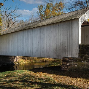 Puede incluir: Un puente cubierto de madera blanca con una base de piedra cruza un pequeño arroyo. El puente está rodeado de árboles con follaje otoñal.