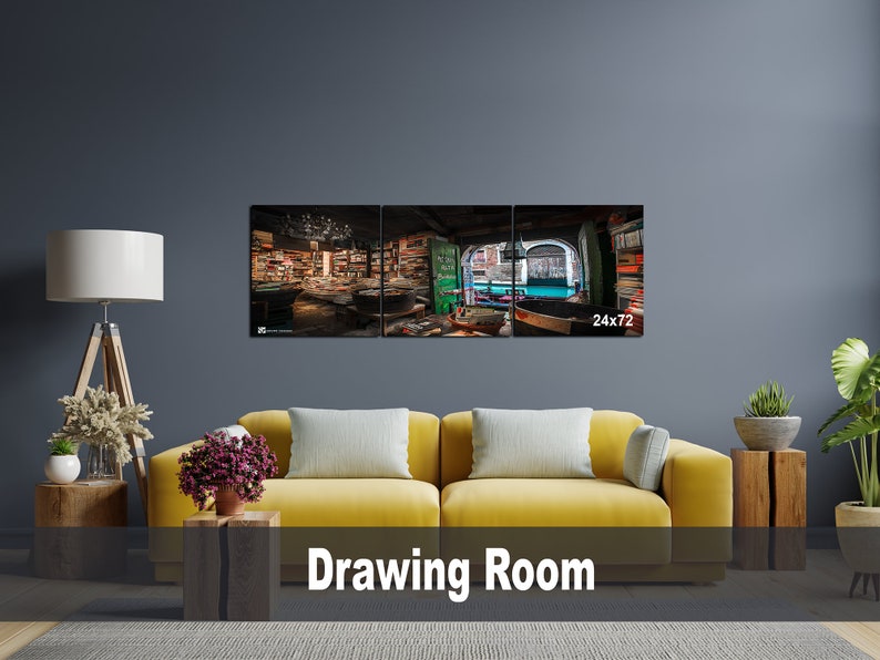 May include: A three-panel canvas print depicting a bookstore interior with a view of a canal. The print is displayed on a wall above a yellow couch in a living room setting. The print measures 24 inches by 72 inches.
