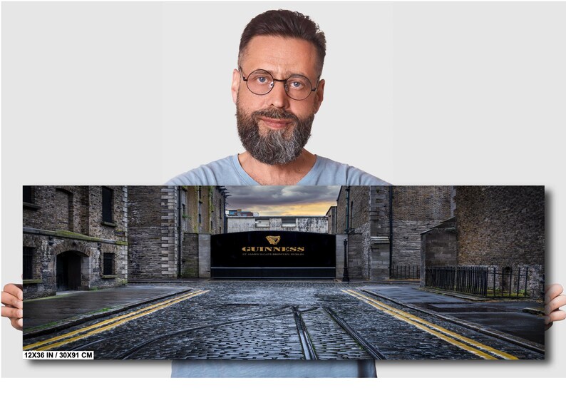 May include: A panoramic photo of a cobblestone street in Dublin, Ireland. The street is lined with old brick buildings. A large sign with the words "GUINNESS" in gold lettering is visible in the distance. The sign is above a large archway. The photo is 12x36 inches.