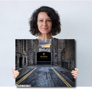 May include: A black and white photograph of a narrow street in Dublin, Ireland. The street is lined with old brick buildings and leads to a large black gate with the word "GUINNESS" in gold lettering. The street is paved with cobblestones and has a yellow stripe down the center.