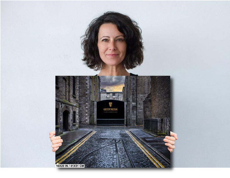 May include: A black and white photograph of a narrow street in Dublin, Ireland. The street is lined with old brick buildings and leads to a large black gate with the word "GUINNESS" in gold lettering. The street is paved with cobblestones and has a yellow stripe down the center.