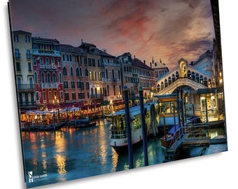 May include: A scenic view of a canal in Venice, Italy at dusk. The canal is lined with colorful buildings and a bridge crosses the water. There are boats docked along the canal and the sky is a vibrant orange and pink.