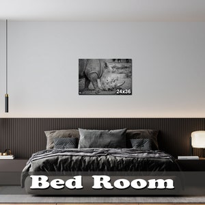 May include: A black and white photograph of a rhinoceros in a grassy field. The photo is framed and hanging on a wall in a modern bedroom with a dark wood headboard and a black and white bedspread. The photo is labeled "24x36".