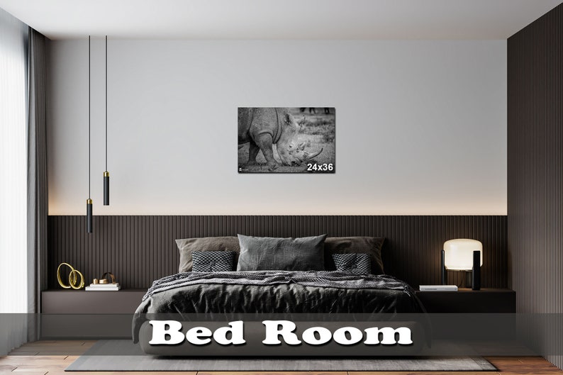 May include: A black and white photograph of a rhinoceros in a grassy field. The photo is framed and hanging on a wall in a modern bedroom with a dark wood headboard and a black and white bedspread. The photo is labeled "24x36".