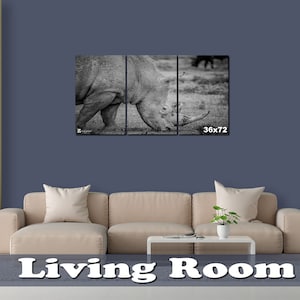 May include: A black and white photo of a rhinoceros in a living room setting. The photo is split into three panels and is framed. The photo is hanging on a wall above a beige couch. The wall is a dark blue color. The couch has three pillows and a coffee table in front of it. The photo is 36 inches by 72 inches.