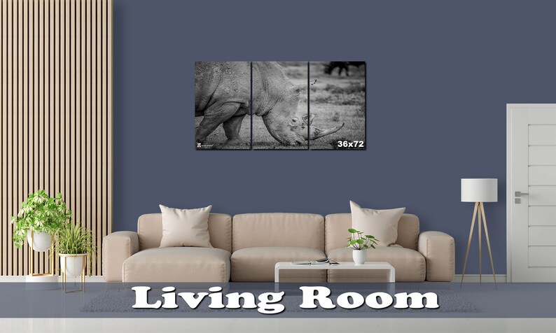 May include: A black and white photo of a rhinoceros in a living room setting. The photo is split into three panels and is framed. The photo is hanging on a wall above a beige couch. The wall is a dark blue color. The couch has three pillows and a coffee table in front of it. The photo is 36 inches by 72 inches.
