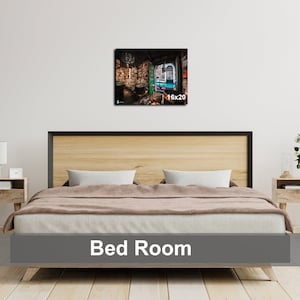 May include: A bedroom with a wooden bed frame and a brown blanket. There is a nightstand with a lamp on each side of the bed. A framed print of a room with bookshelves and a window is hanging on the wall above the bed. The print is labeled "16x20".
