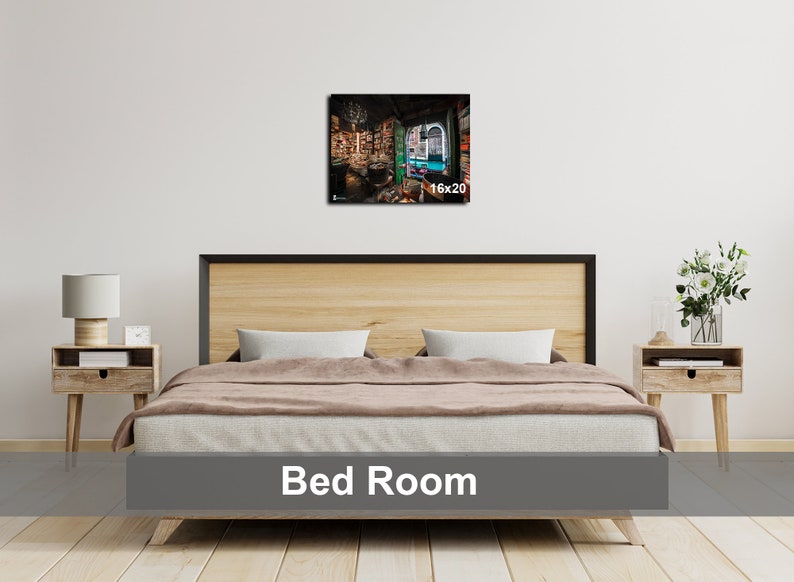 May include: A bedroom with a wooden bed frame and a brown blanket. There is a nightstand with a lamp on each side of the bed. A framed print of a room with bookshelves and a window is hanging on the wall above the bed. The print is labeled "16x20".