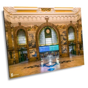 Puede incluir: El interior de una gran estación de tren adornada con un tablero de visualización digital azul que muestra los horarios de los trenes. La estación tiene un techo abovedado alto con molduras decorativas y paredes de azulejos. La palabra "MINHO" está escrita en letras doradas sobre la entrada.