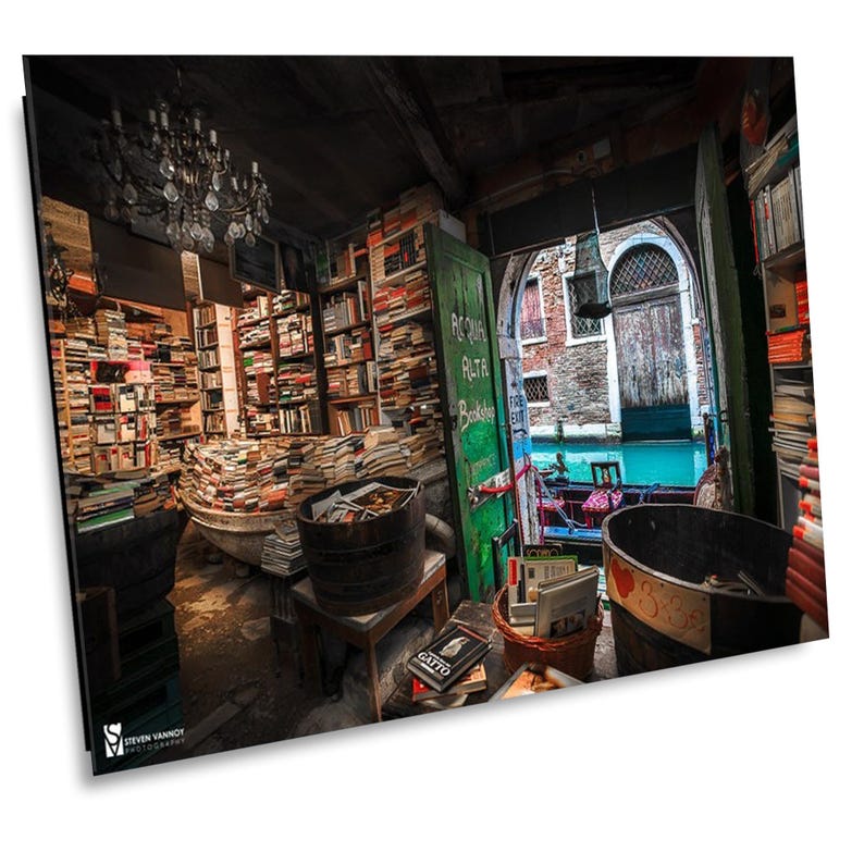 May include: A bookstore in Venice, Italy, with bookshelves filled with books. The store is open to the outside, with a view of a canal. The sign above the doorway reads "Piccola Alta Libreria".