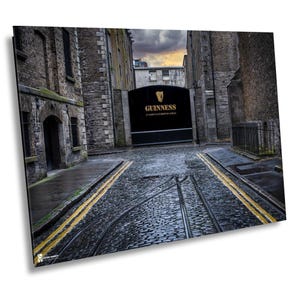May include: A narrow cobblestone street lined with old stone buildings leads to a large black gate with the words "GUINNESS" in gold lettering. The gate is at the end of the street, and the street is wet from rain.