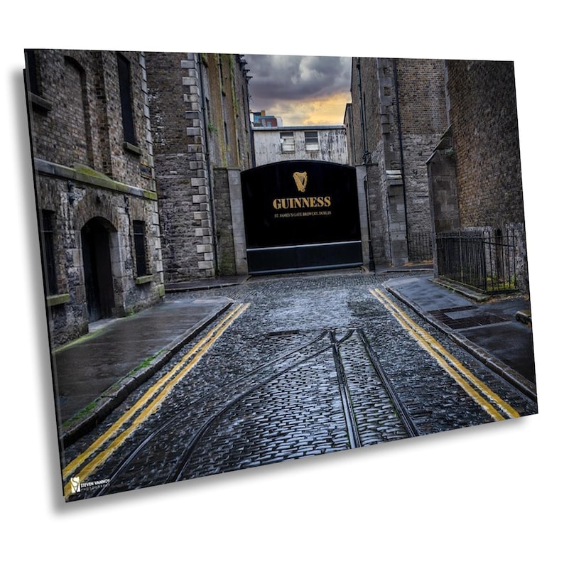 May include: A narrow cobblestone street lined with old stone buildings leads to a large black gate with the words "GUINNESS" in gold lettering. The gate is at the end of the street, and the street is wet from rain.