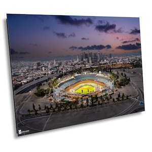 Pode incluir: Vista aérea de um estádio de beisebol ao entardecer, com o horizonte da cidade ao fundo. O estádio está brilhantemente iluminado, contrastando com o céu escuro. A imagem captura a arquitetura do estádio e a paisagem urbana.