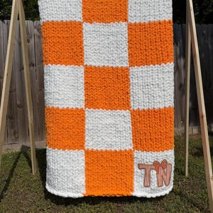 May include: A large, handmade, checkerboard-patterned blanket in orange and white. The blanket is displayed outdoors, hanging from a wooden frame. The letters "TN" are embroidered on the bottom right corner.
