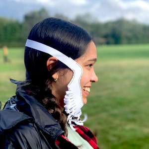 Op de afbeelding: Een persoon draagt ​​een witte papieren kostuummal met een gekarteld randontwerp. De mal wordt over het hoofd van de persoon gedragen en bedekt het gezicht. De persoon glimlacht en kijkt naar rechts. De achtergrond is een groen grasveld.