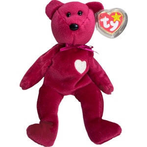 May include: A plush pink beanie baby bear with a white heart on its chest and a pink ribbon around its neck. The bear has black eyes and a black nose. The bear is sitting on a white background. The Ty Beanie Baby tag is attached to the bear's ear.