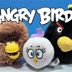 Op de afbeelding: Drie pluche Angry Birds-personages: een bruine vogel met een gele snavel, een witte vogel met een paarse kuif en een zwarte vogel met rode wenkbrauwen. Het Angry Birds-logo wordt boven de personages weergegeven.
