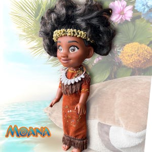 Side profile of the Moana doll standing on the shoreline with tropical foliage in the background. Her curly hair is adorned with a real ocean snail shell headband.