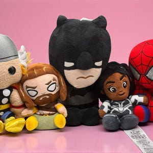 May include: A group of plush superhero toys, featuring Thor, Batman, Spider-Man, and Black Panther, set against a pink backdrop. The soft toys are brightly coloured and have cartoon-like characteristics. Each character is instantly recognisable.