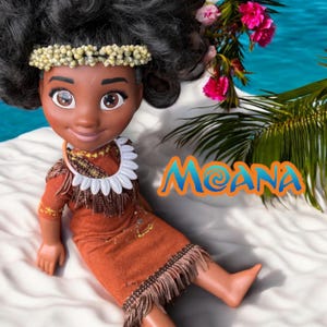 Moana doll sitting on soft white sand with turquoise water in the background. She wears a handmade terracotta dress with fringe and a real shell headband. The Moana logo is in the corner.