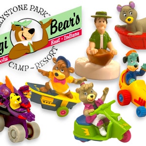 Pode incluir: Um conjunto de sete brinquedos de plástico com tema Yogi Bear e Jellystone Park. Os brinquedos incluem Yogi Bear em um skate, Boo Boo Bear em um scooter, Cindy Bear em um carro de corrida, Huckleberry Hound em um barco, Ranger Smith em um barco, um caminhão monstro roxo e preto, e um urso marrom com uma fralda em um barco vermelho. Os brinquedos estão todos em várias poses e têm cores brilhantes.