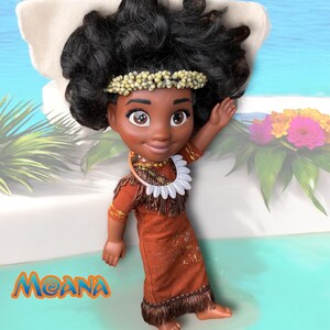 Moana doll leaning on a cushion by the water, one arm raised. She wears a custom tribal-style dress and shell necklace, with her signature textured curls.