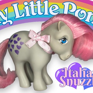 May include: A gray My Little Pony with pink hair and a purple heart pattern on its rear. The pony has a pink bow around its neck and blue eyes. The pony is standing on a white background with a rainbow and the words "My Little Pony" above it. The words "Italian Snuggle" are printed in a purple heart shape below the pony.